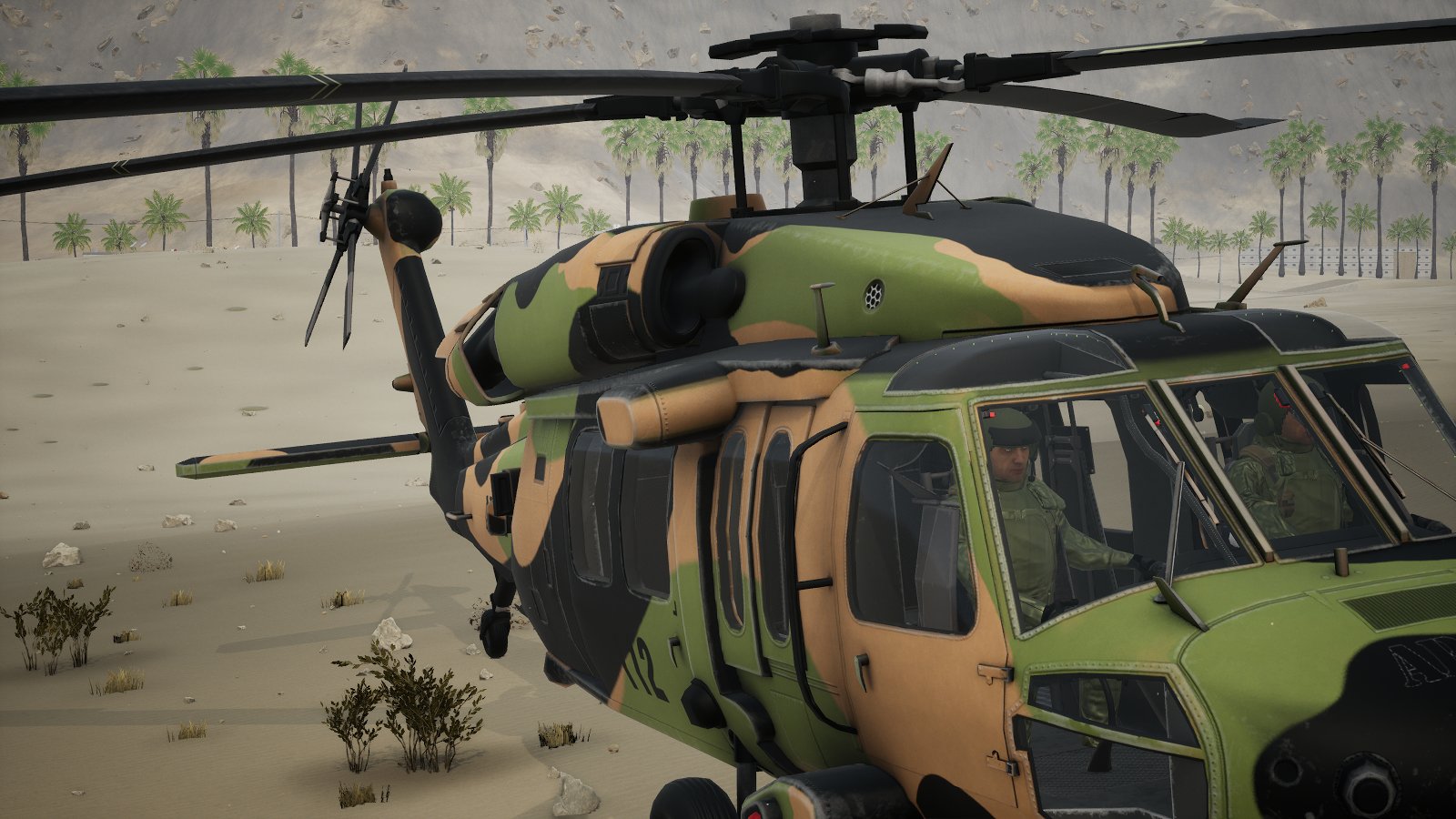 A Black Hawk parked on the sand at Khasab, seen from the front quarter: both pilots in sage flight suits, vests and aircrew helmets with boom mics behind the windscreen.